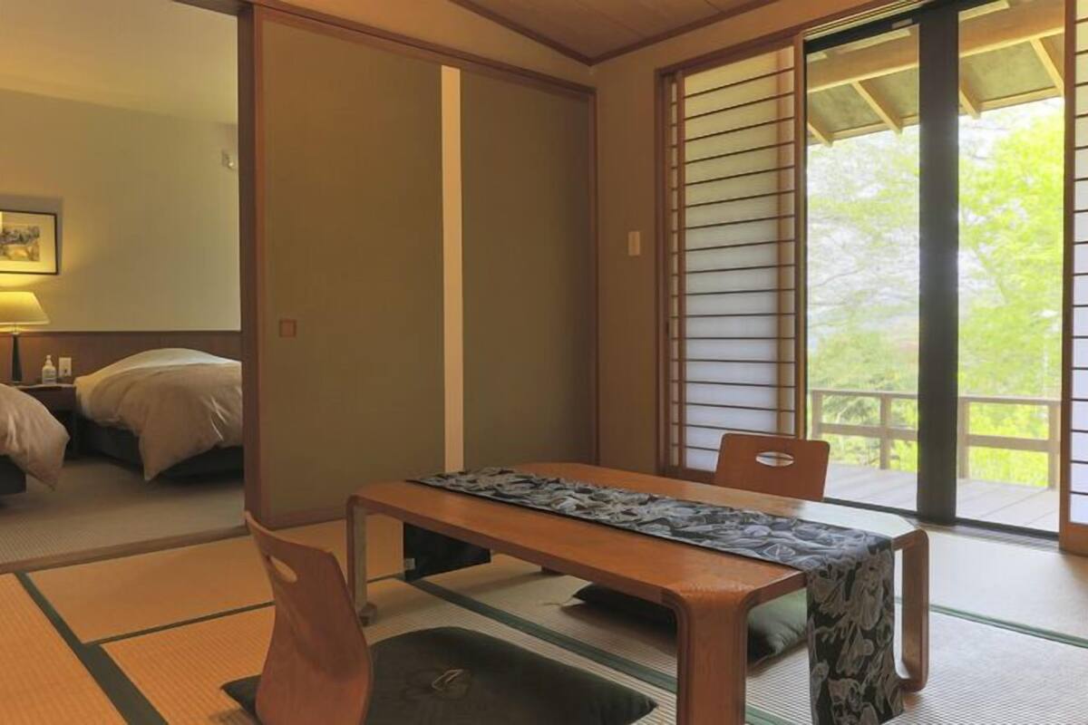 A traditional Japanese room with tatami flooring, a low table with a patterned tablecloth, two beds, and a sliding door leading to an outdoor area.