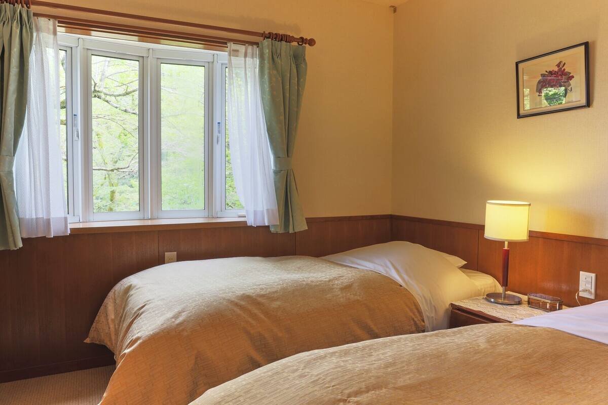 A room with two beds, a window with curtains, a bedside lamp, and a framed picture on the wall.