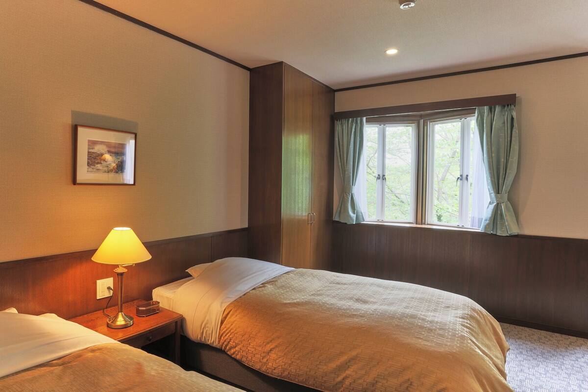 A hotel room with two beds, a bedside lamp, a window with curtains, and a framed picture on the wall.