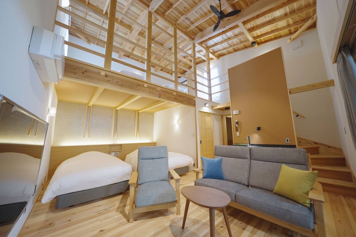 A modern interior with a high ceiling, wooden beams, a sofa, a chair, a small table, and a bed.