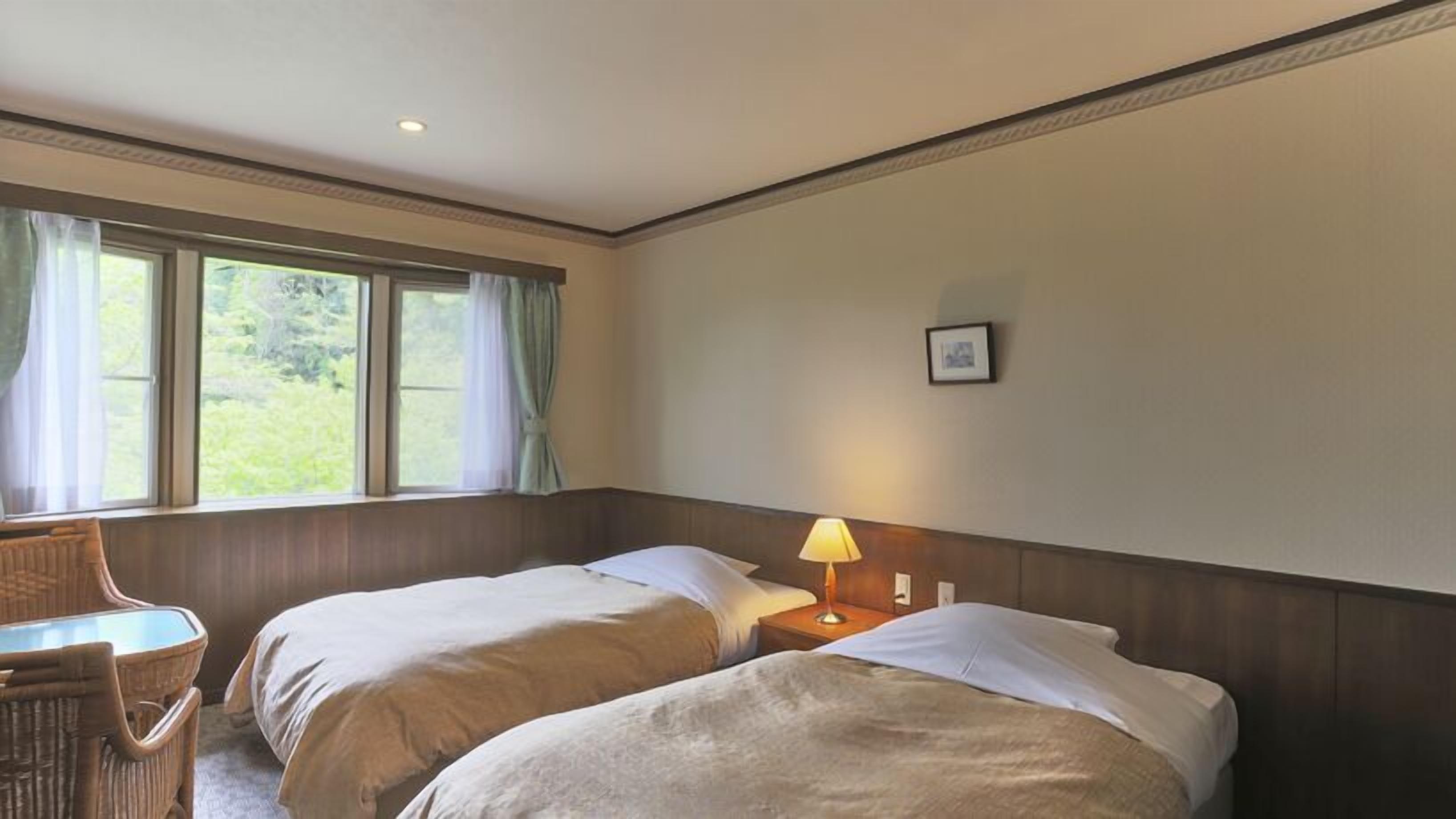 Habitació d'hotel amb dos llits, una taula petita, una cadira, una làmpada i una finestra amb cortines.