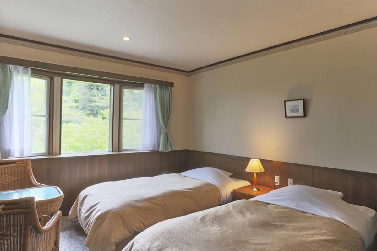 A hotel room with two beds, a small table, a chair, a lamp, and a window with curtains.