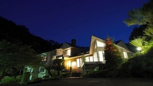 A large, two-story house with a prominent chimney, surrounded by lush greenery and a well-lit driveway. - Mastlamp (Ito)