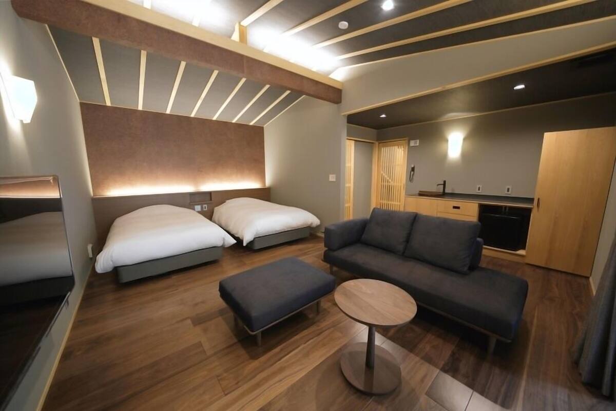 A modern hotel room with a sofa, two beds, a small table, and a kitchenette.