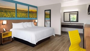 Une chambre d’hôtel avec un grand lit, une table de chevet avec une lampe, un miroir, une salle de bain avec un lavabo et une chaise jaune.