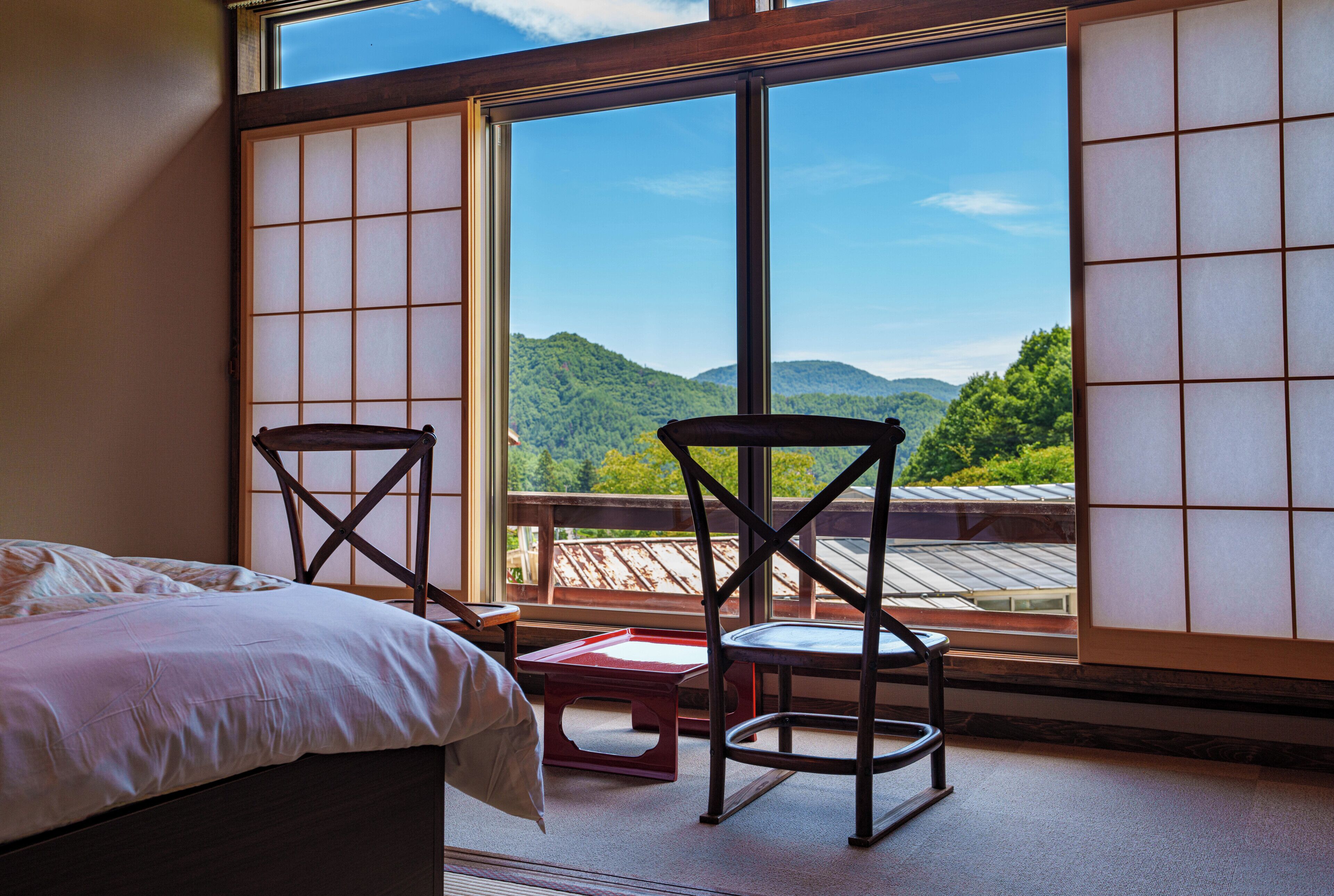 Habitación con cama, dos sillas, una mesita y un amplio ventanal con vistas a un paisaje montañoso.