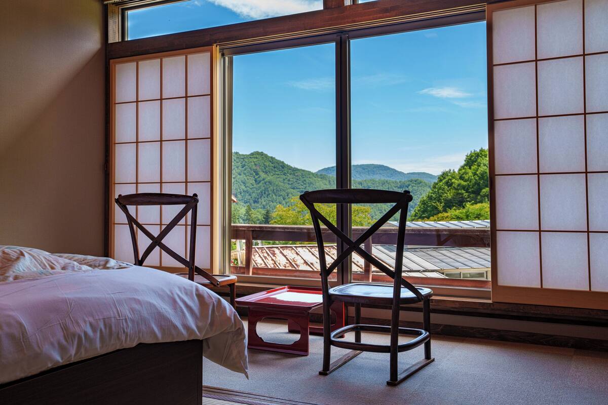 A room with a bed, two chairs, a small table, and a large window offering a view of a mountain landscape.