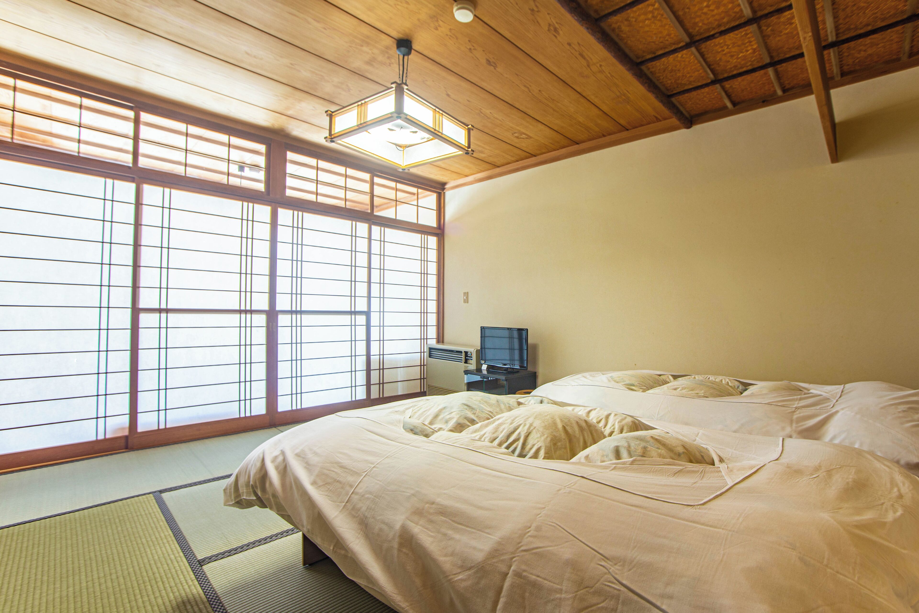Una habitación japonesa tradicional con suelo de tatami, una cama con ropa de cama blanca, un televisor y un ventilador de techo.