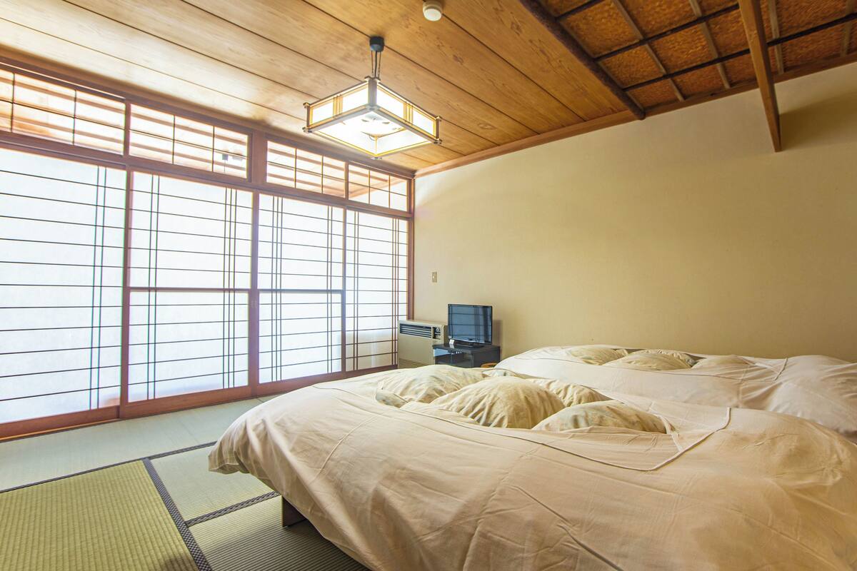 A traditional Japanese room with tatami flooring, a bed with white bedding, a television, and a ceiling fan.