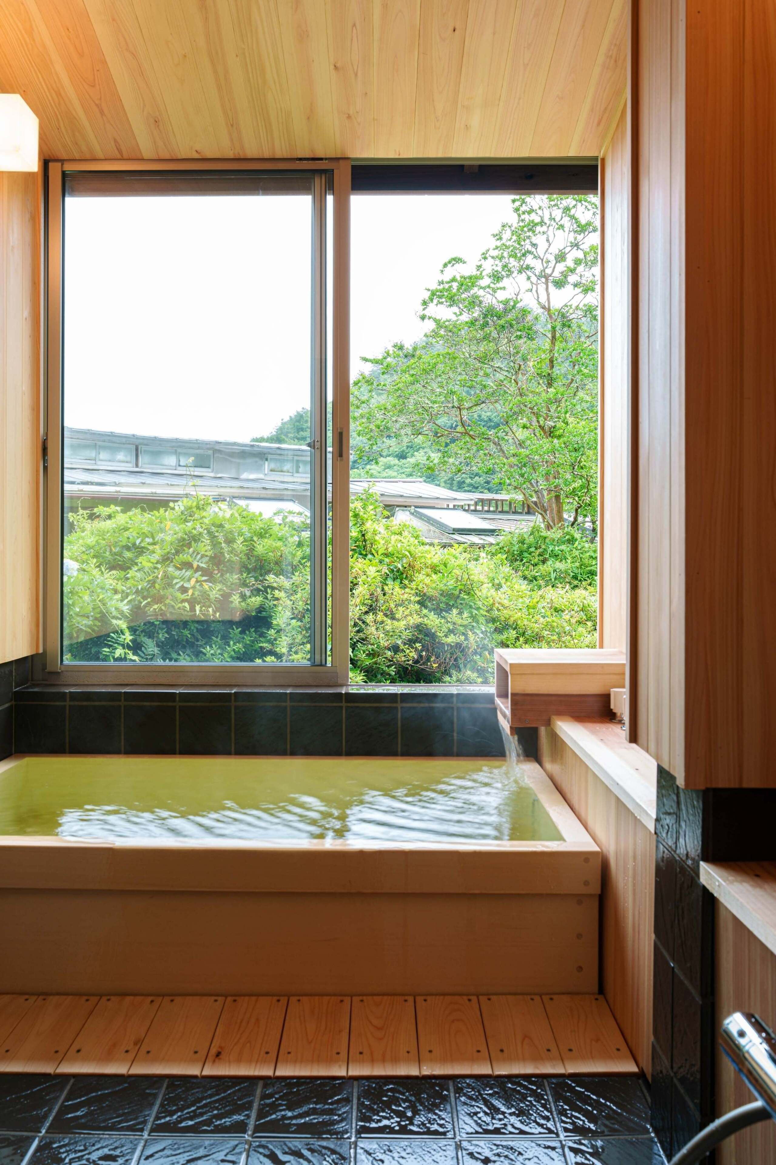A wooden bathtub with a view of greenery and a building outside.