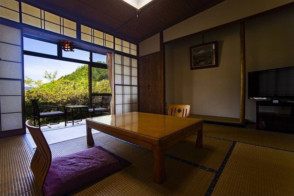 A traditional Japanese room with tatami flooring, a wooden table, chairs, a television, and a view of greenery outside.