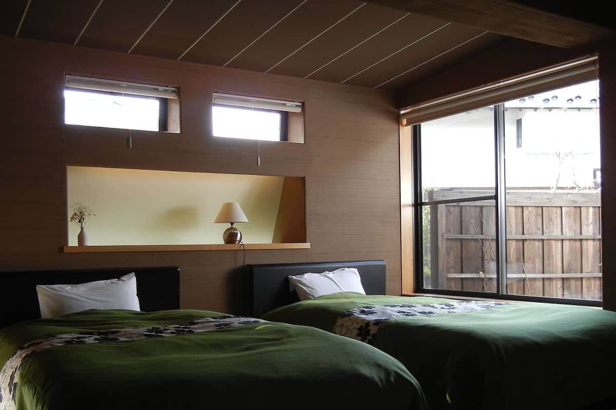A bedroom with two beds, a nightstand, a lamp, and a window with a view of a wooden fence.