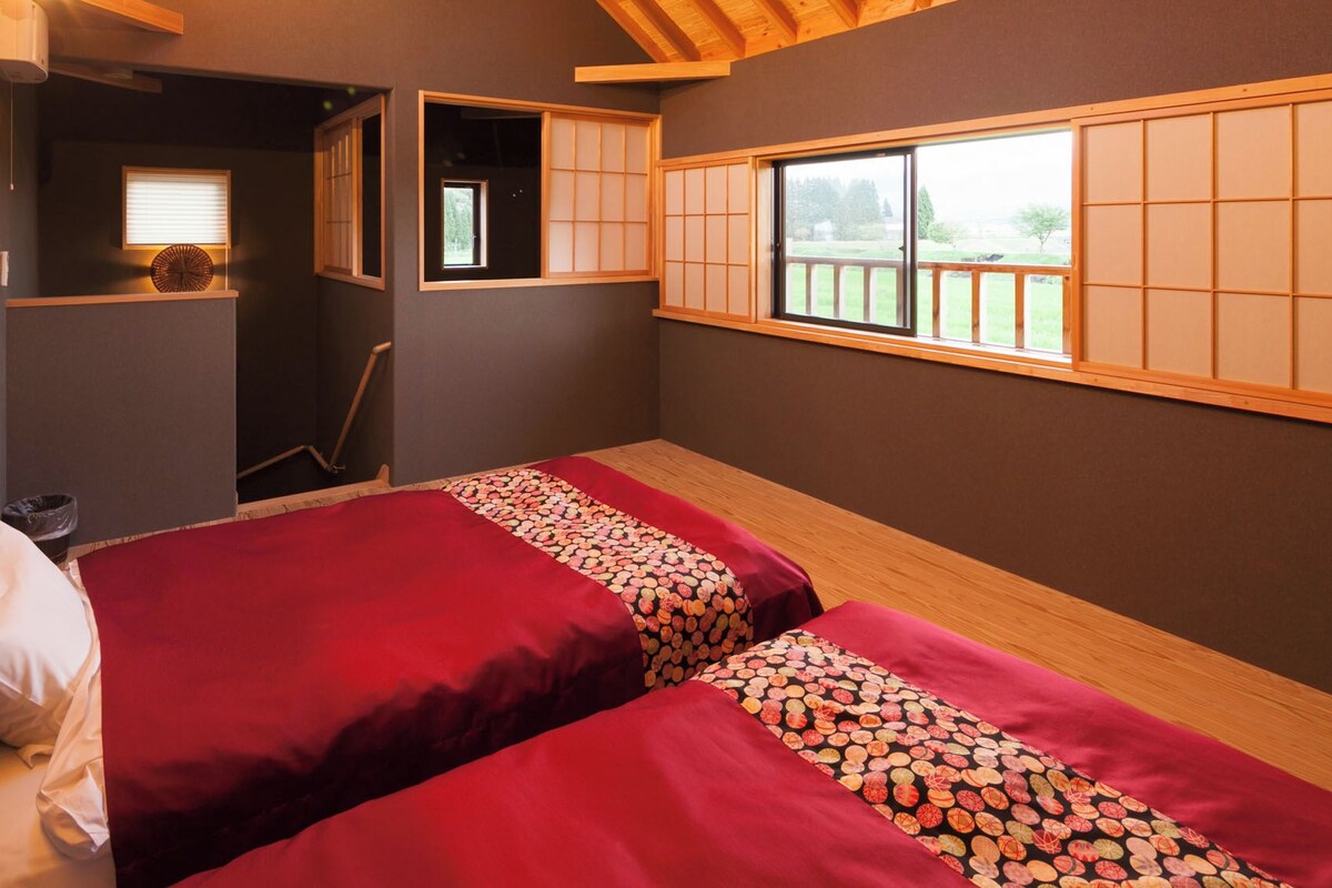 A room with two beds, a window with a view, and a small shelf.