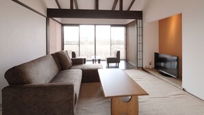 A modern living room with a sectional sofa, a wooden coffee table, a TV, and a sliding glass door leading to an outdoor area. - Takanosho (Aso)