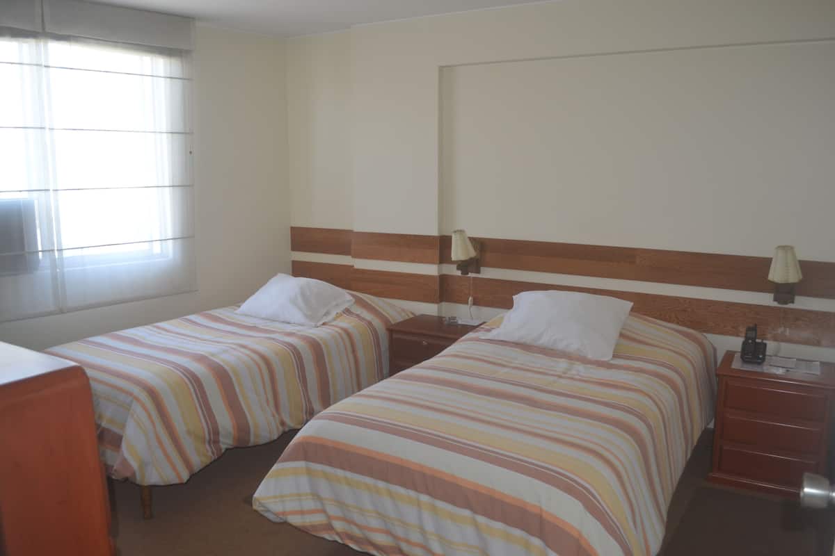 Quarto de hotel com duas camas, mesa de cabeceira, abajur e janela com persianas.