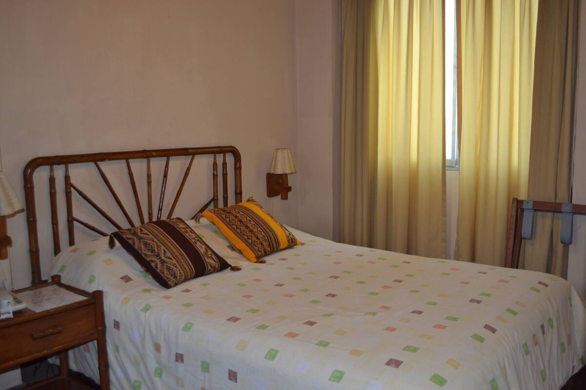 Quarto com cabeceira de madeira, mesa de cabeceira com abajur, cama com almofadas estampadas e janela com cortinas.