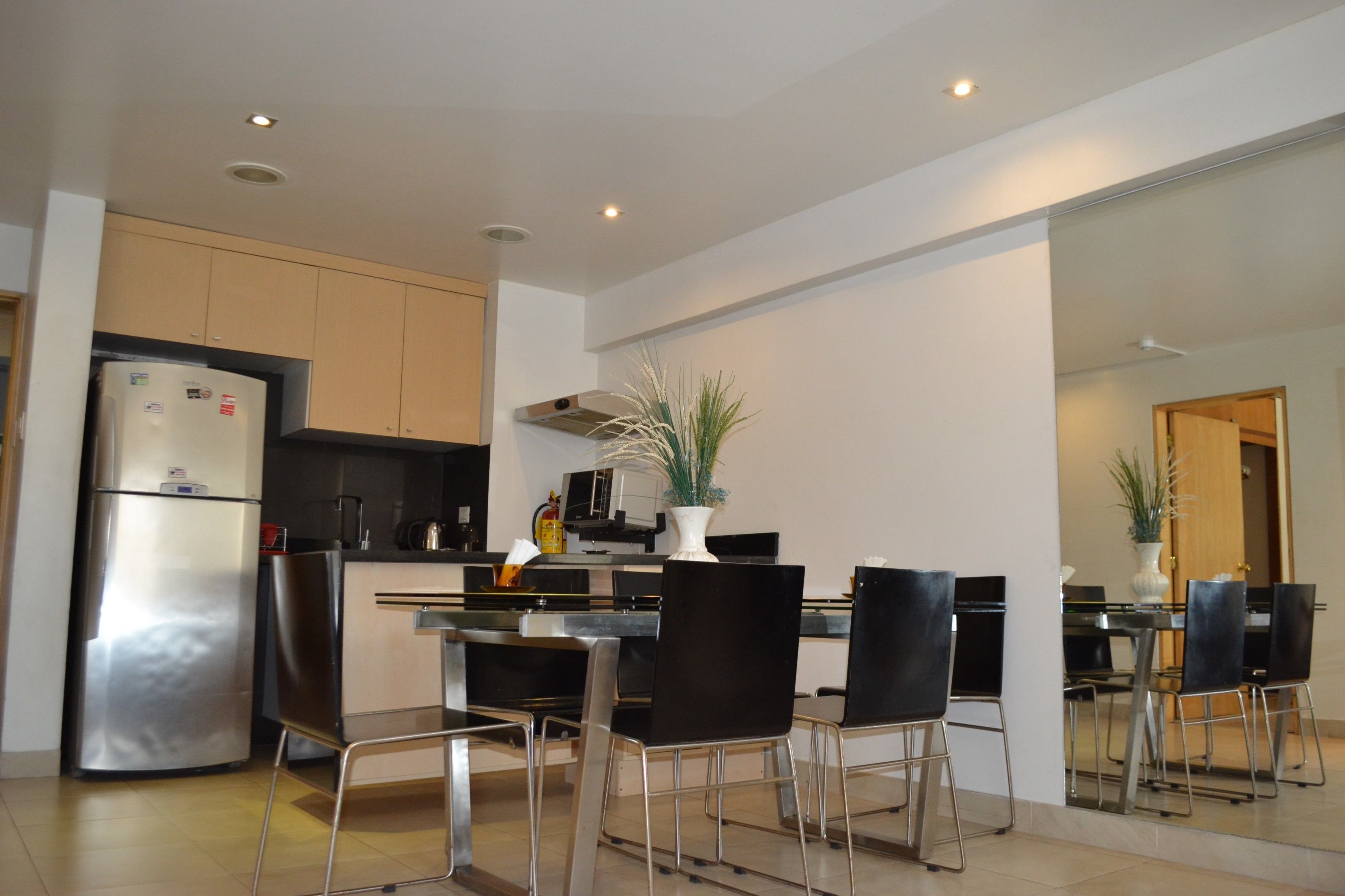A modern kitchen with a stainless steel refrigerator, black chairs around a dining table, and a large mirror reflecting the space.