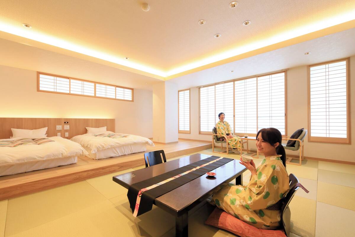 A room with a long table, two beds, and large windows.