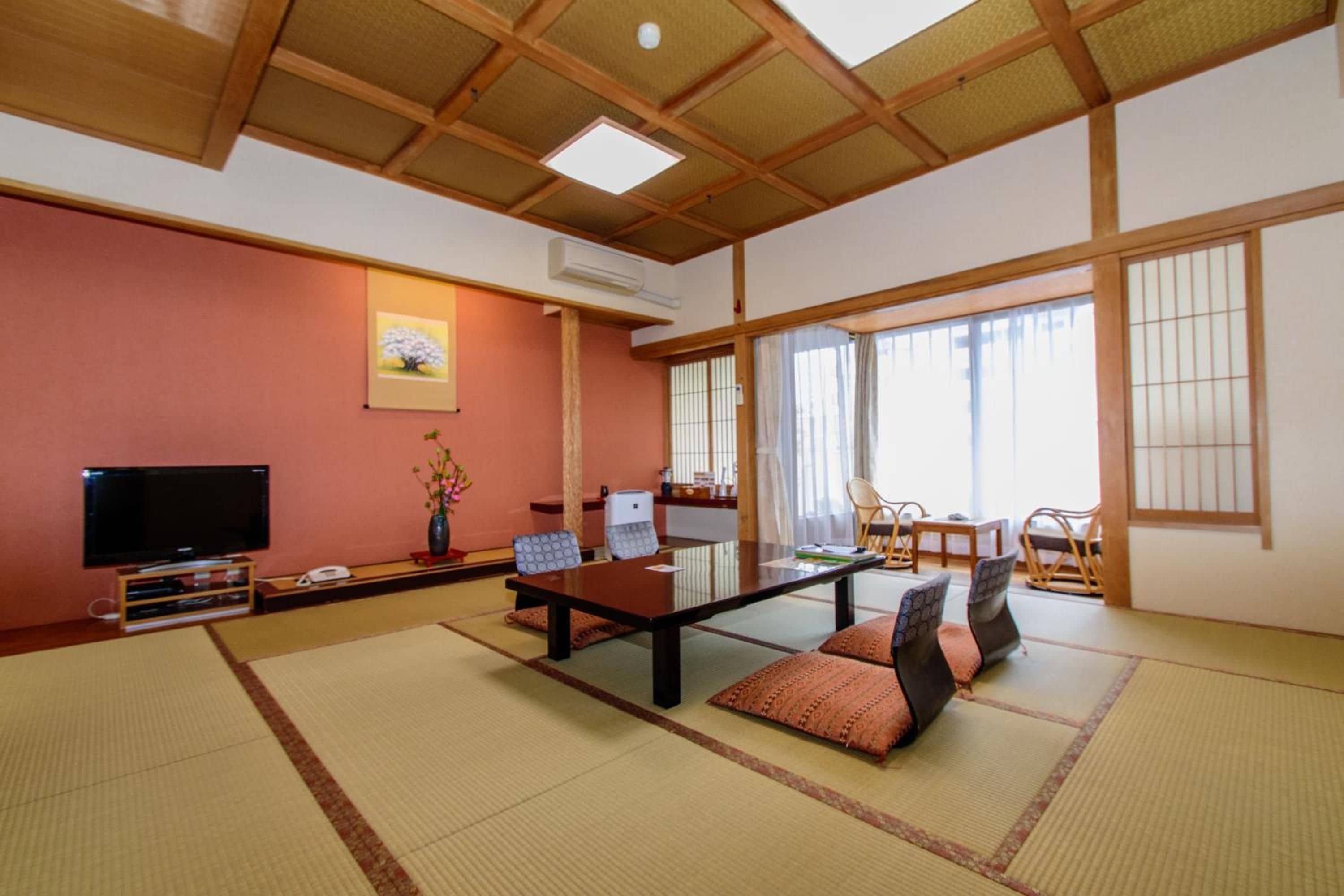 Ein traditioneller japanischer Raum mit Tatami-Boden, einem niedrigen Tisch mit Kissen, einem Fernseher und einem großen Fenster mit Vorhängen.