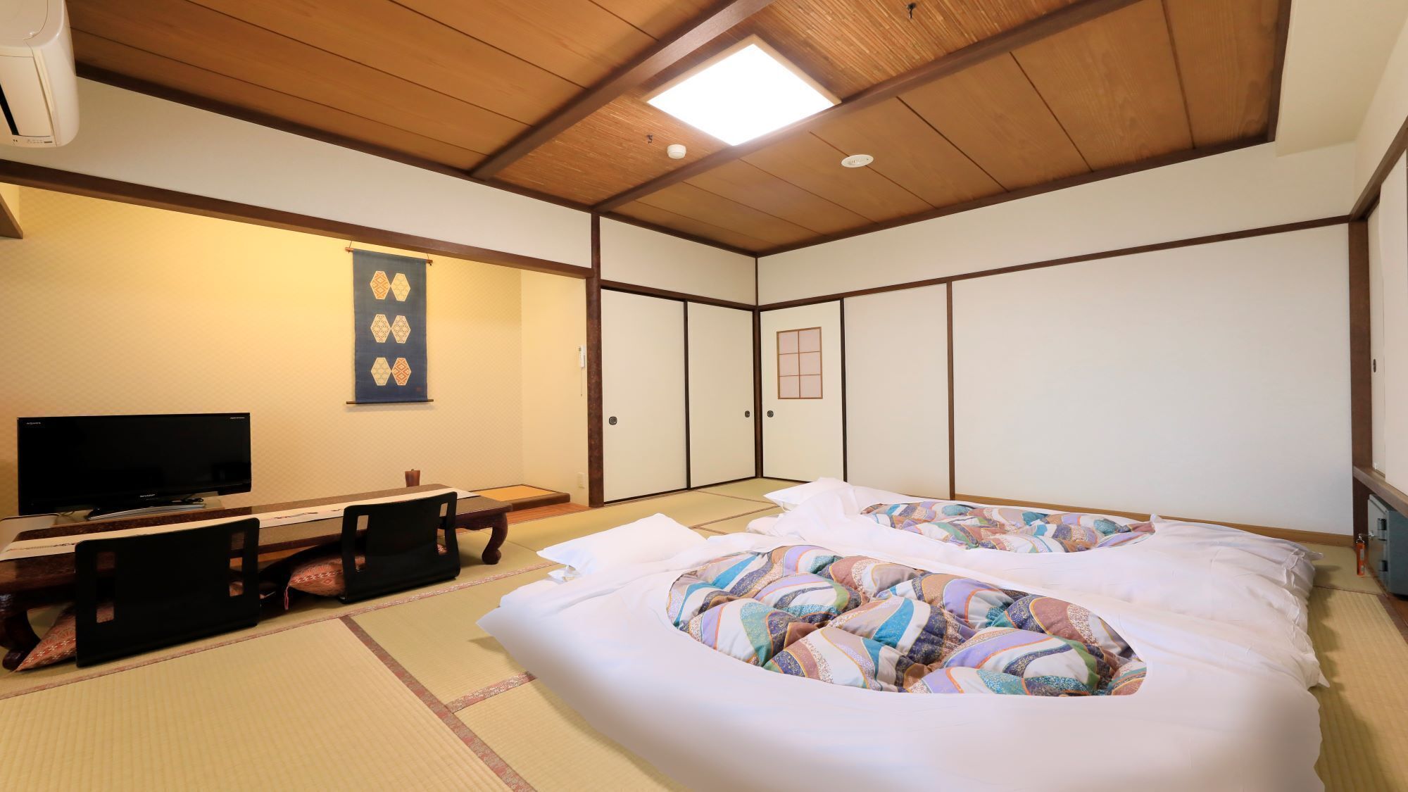 Una habitación japonesa tradicional con suelo de tatami, una cama con ropa de cama colorida, un televisor, un escritorio con una silla y puertas correderas.