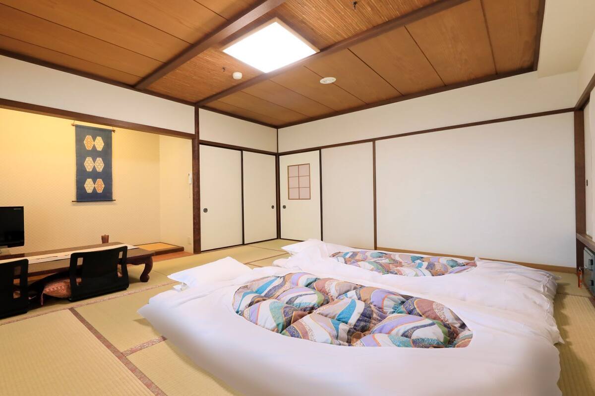 A traditional Japanese room with tatami flooring, a bed with colorful bedding, a television, a desk with a chair, and sliding doors.