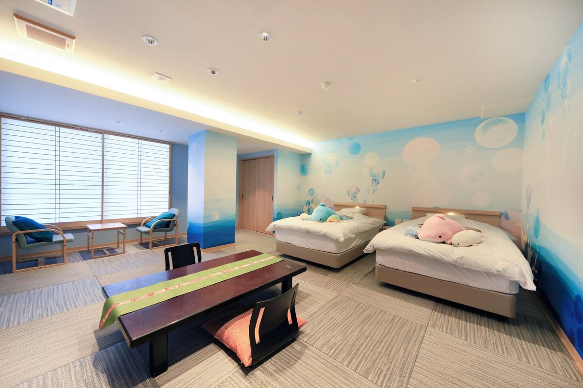 A hotel room with two beds, a desk, chairs, and a large window with blinds.