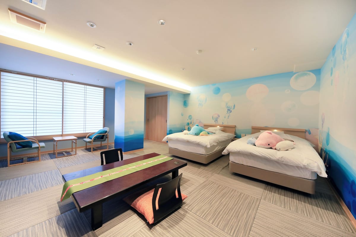 Habitación de hotel con dos camas, un escritorio, sillas y un ventanal con persianas.