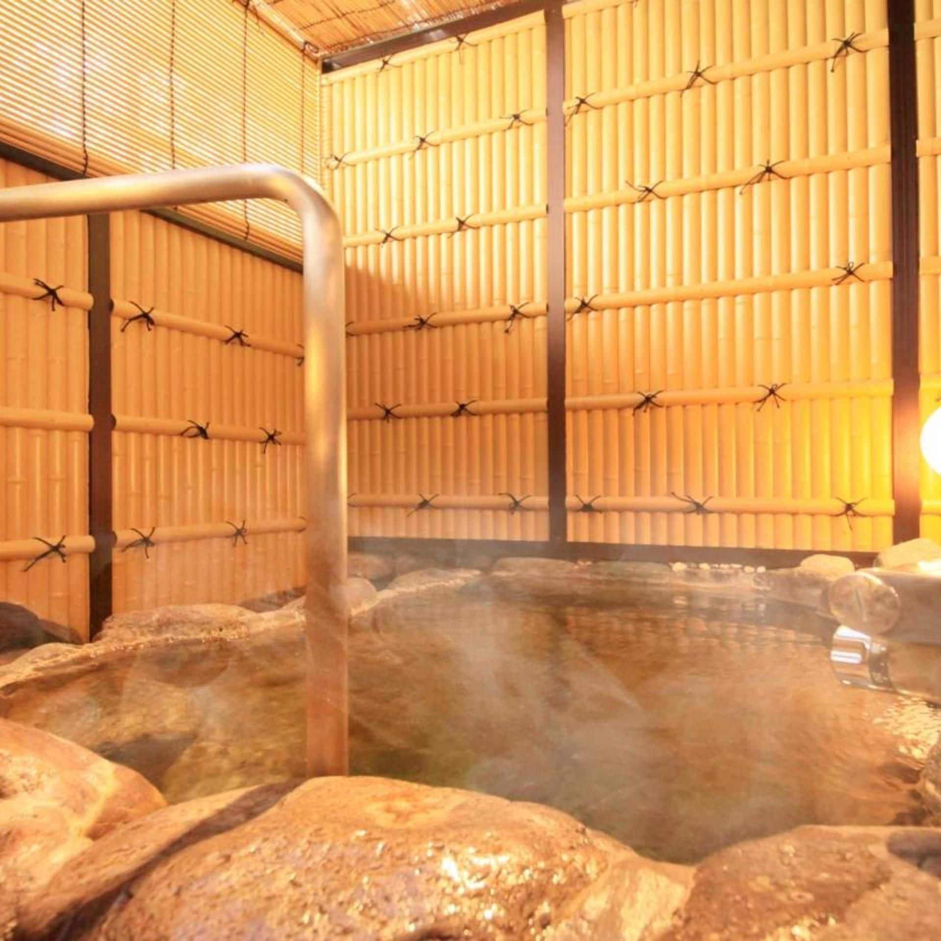 A hot spring area with a metal ladder leading into a stone-lined pool, surrounded by bamboo walls.