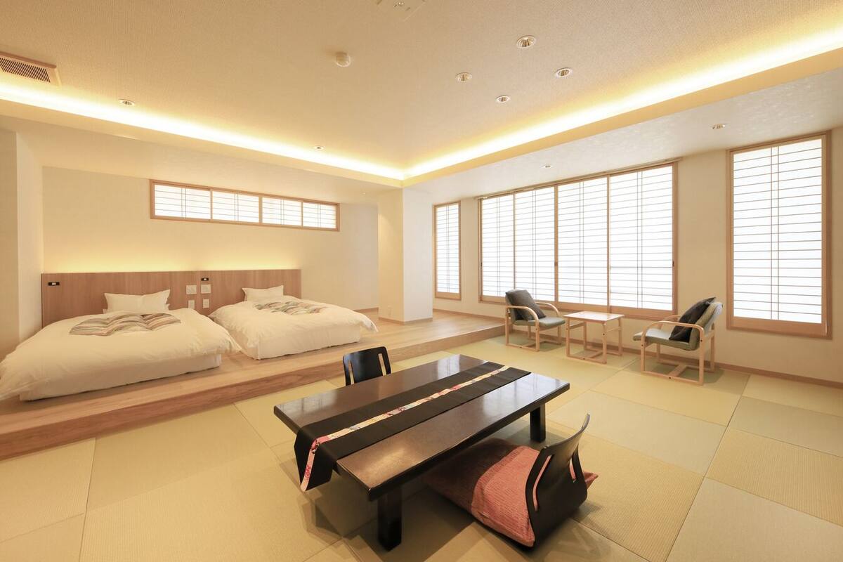 A hotel room with a bed, two chairs, a table, and large windows with blinds.