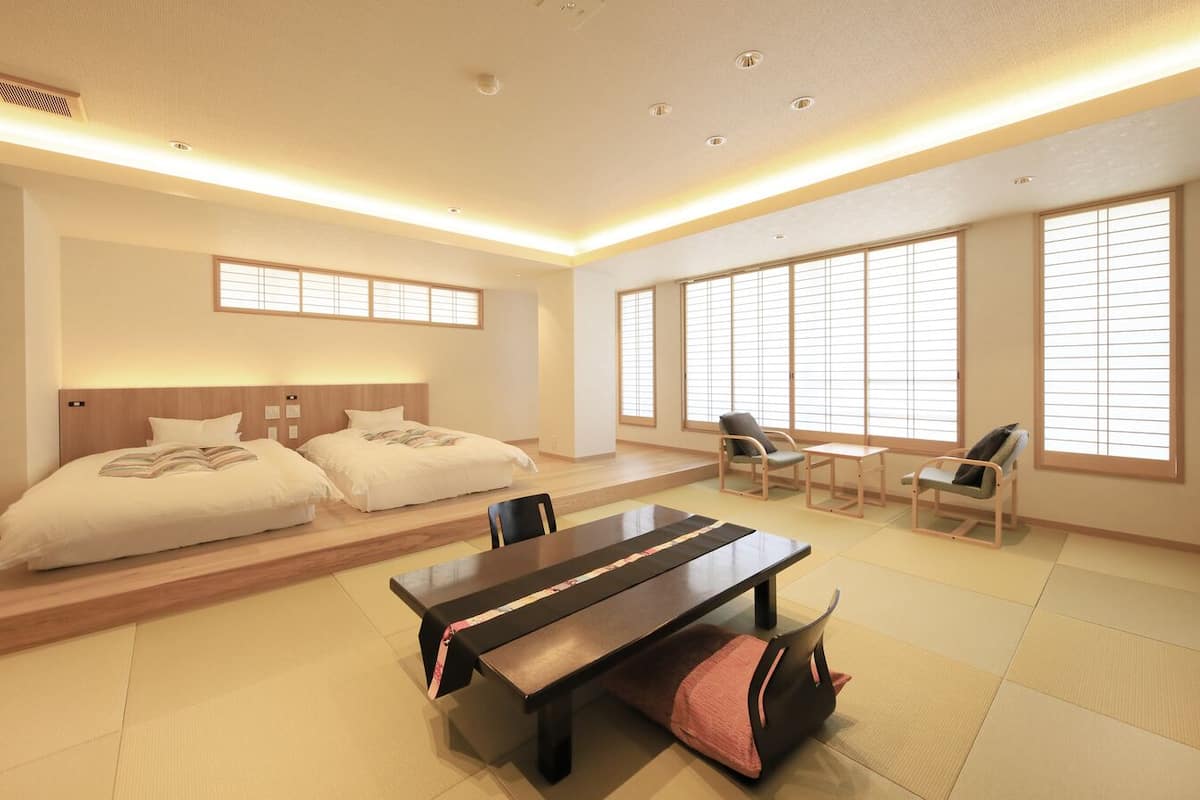 Una habitación de hotel con una cama, dos sillones, una mesa y ventanas amplias con persianas.