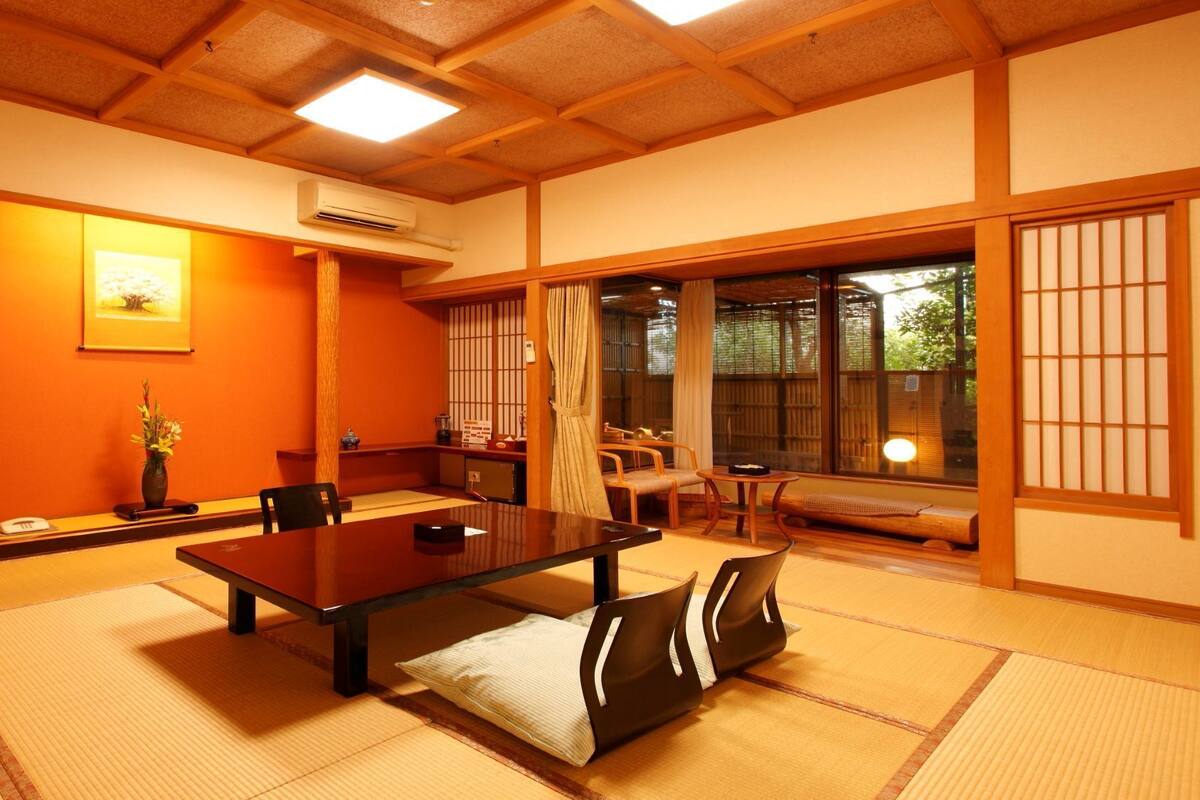A traditional Japanese room with tatami flooring, a low table, chairs, a television, and a large window with sliding doors.