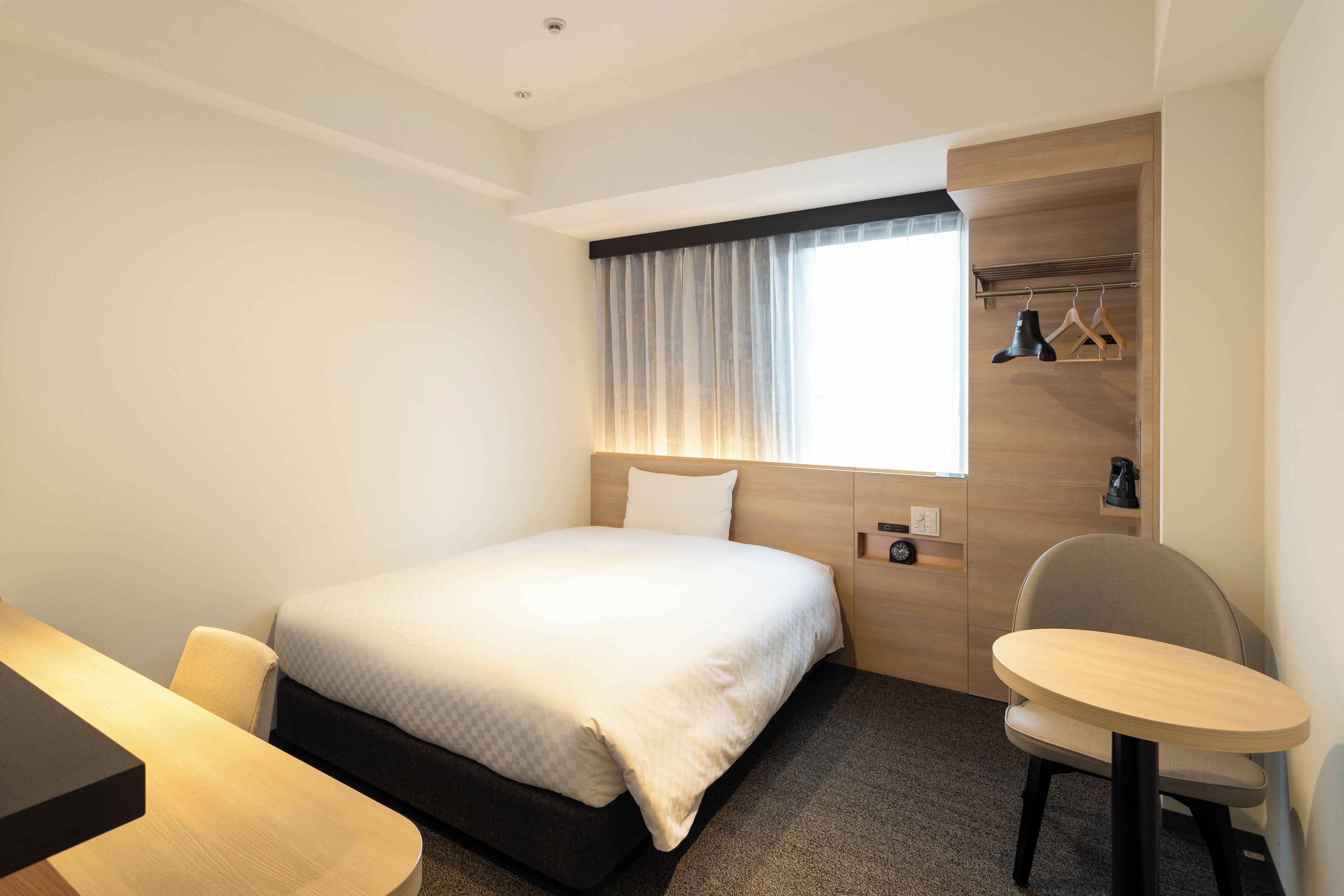 A hotel room with a bed, a desk, a chair, a small table, a coat rack, and a window with curtains.
