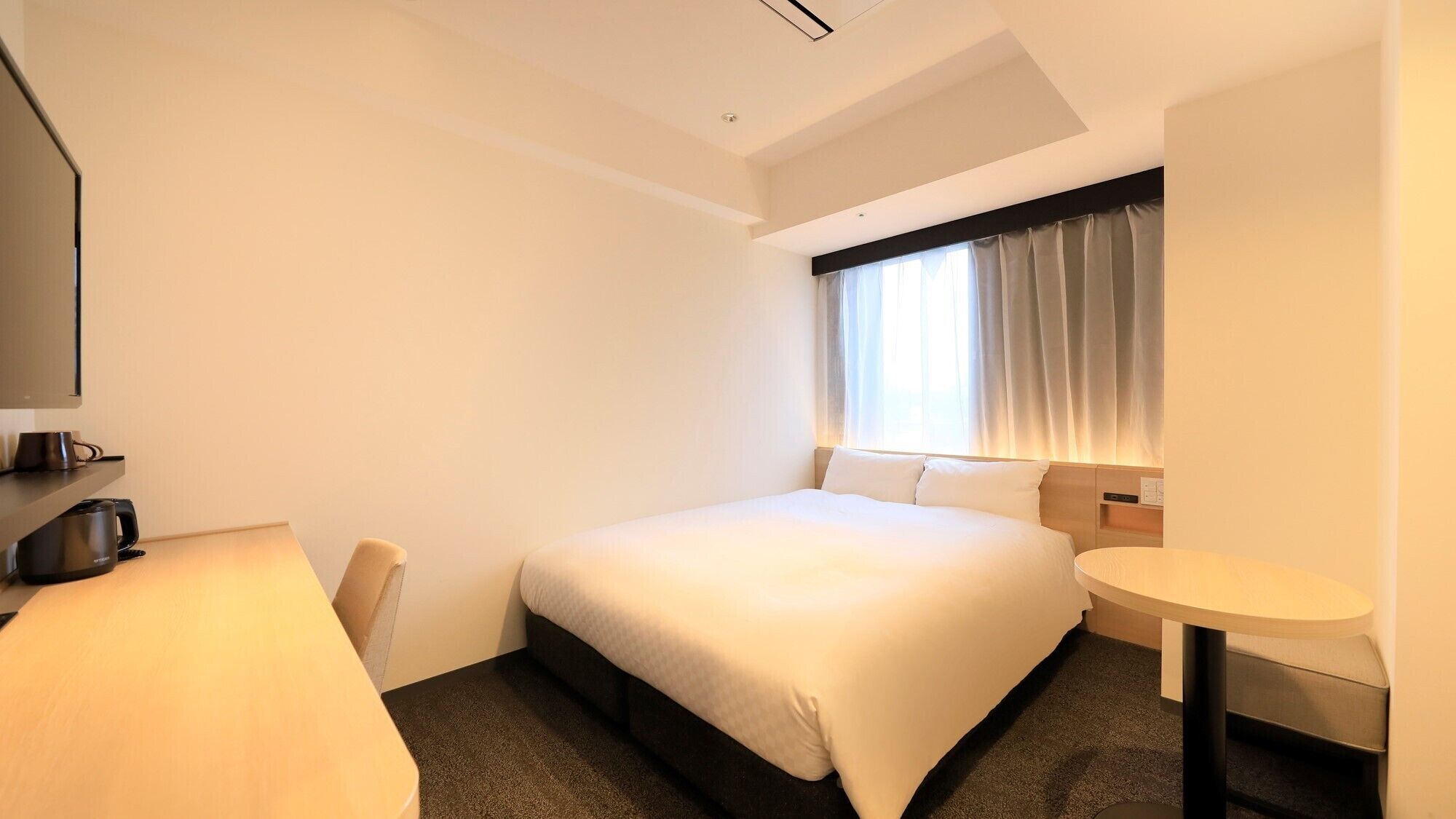 Habitación de hotel con cama, escritorio, silla, una mesita y ventana con cortinas.
