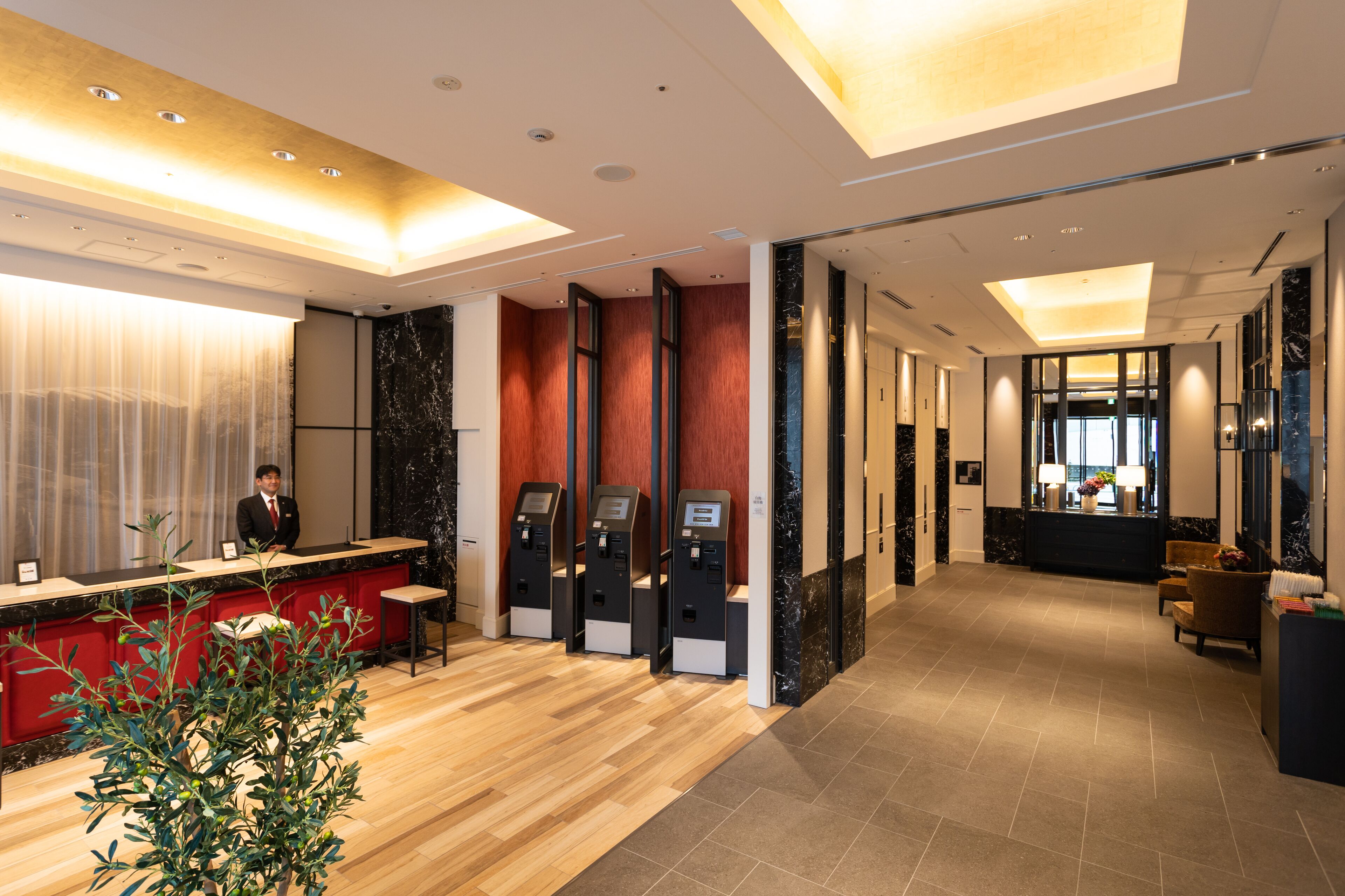A modern hotel lobby with a reception desk, self-service kiosks, a reception area, and a seating area with a plant.