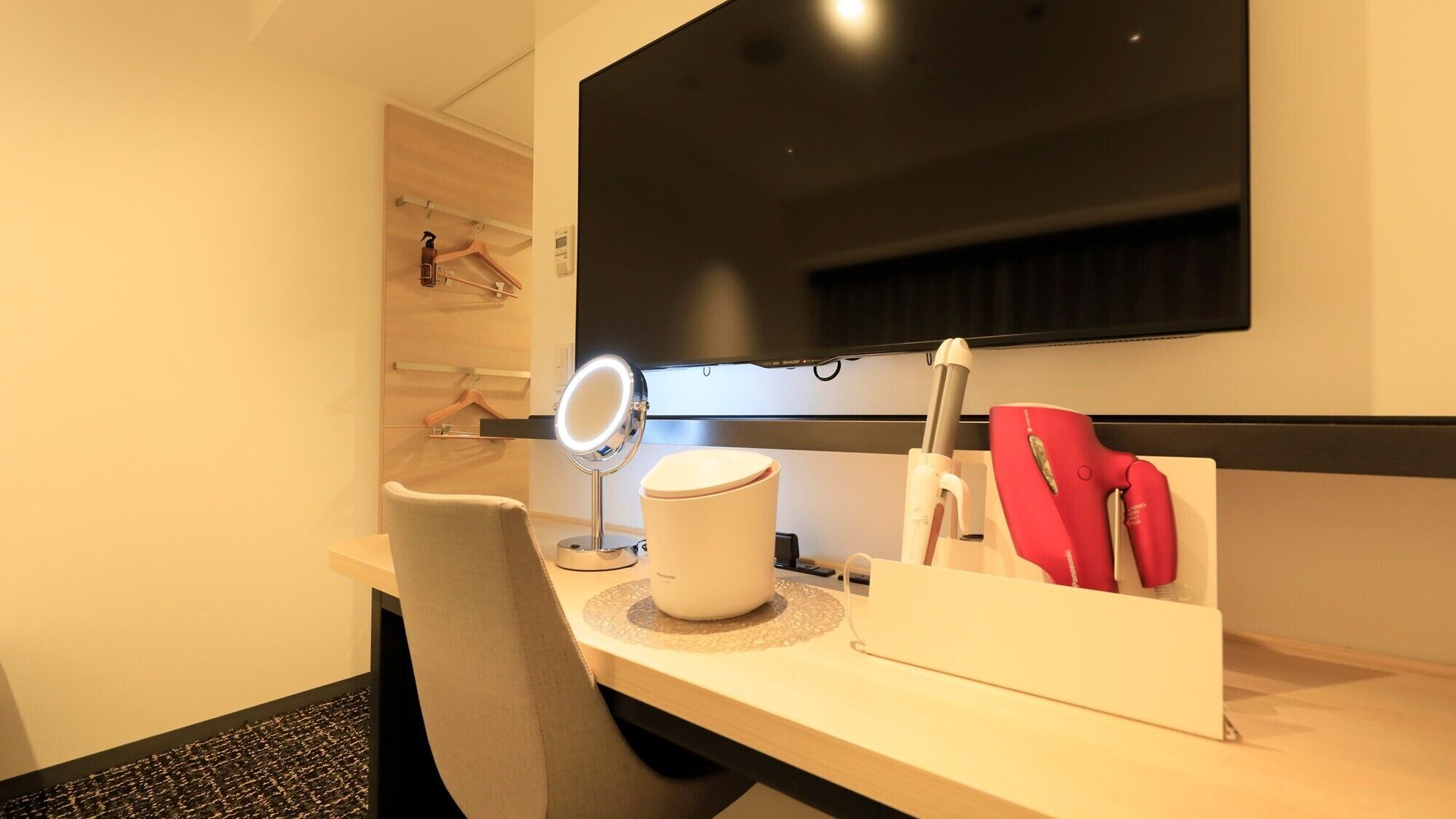 Habitación de hotel con escritorio, una silla, tocador con espejo, secador de pelo y televisor de pantalla plana montado en la pared.