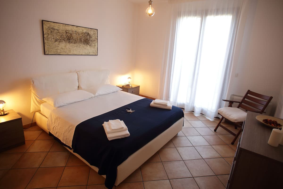 A neatly made bed with a dark blue bedspread and white pillows, a bedside lamp, a wooden nightstand, a chair, and a dining table with a plate of fruit.