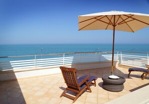 Una terraza con una silla de madera, un asiento acolchado, un taburete de mimbre y un paraguas grande.