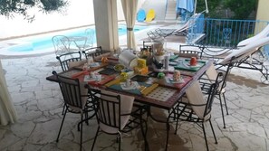 An outdoor dining area with a table set for a meal, surrounded by chairs, and a pool in the background. - B&B Rita and Family (Loceri)
