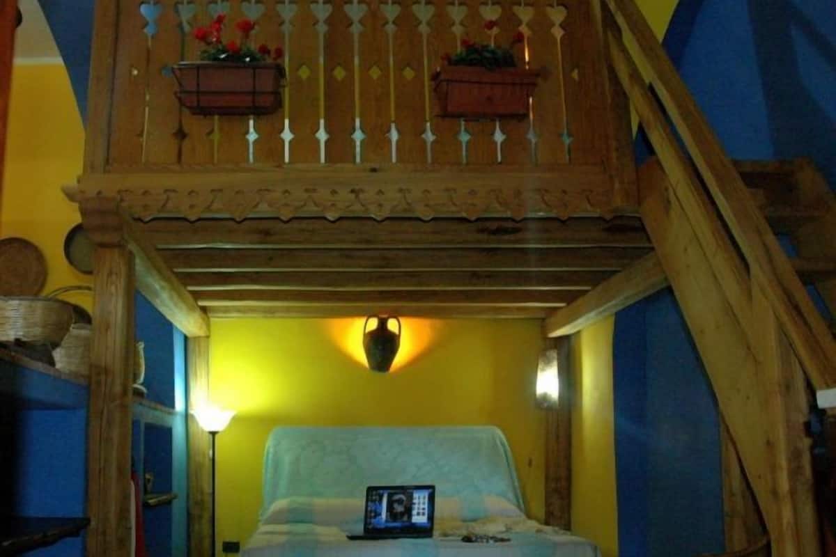 A room with a bed, a laptop on the bed, a wooden balcony with flower baskets, and a lamp on the wall.