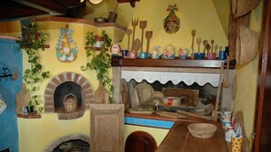 A rustic kitchen with a brick oven, wooden shelves with kitchen utensils, and a wooden table. - B&B Rita and Family (Loceri)