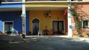 Un edificio con fachada azul y amarilla, un acceso cubierto con una mesa y sillas, y una persona de pie cerca de la entrada.