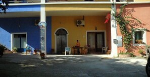 A building with a blue and yellow facade, a covered entrance with a table and chairs, and a person standing near the entrance. - B&B Rita and Family (Loceri)