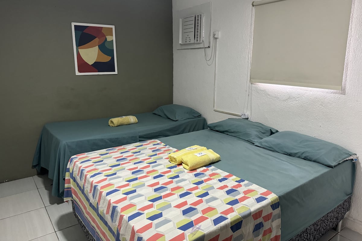 A room with two single beds, a colorful quilt, a painting, and a window with blinds.