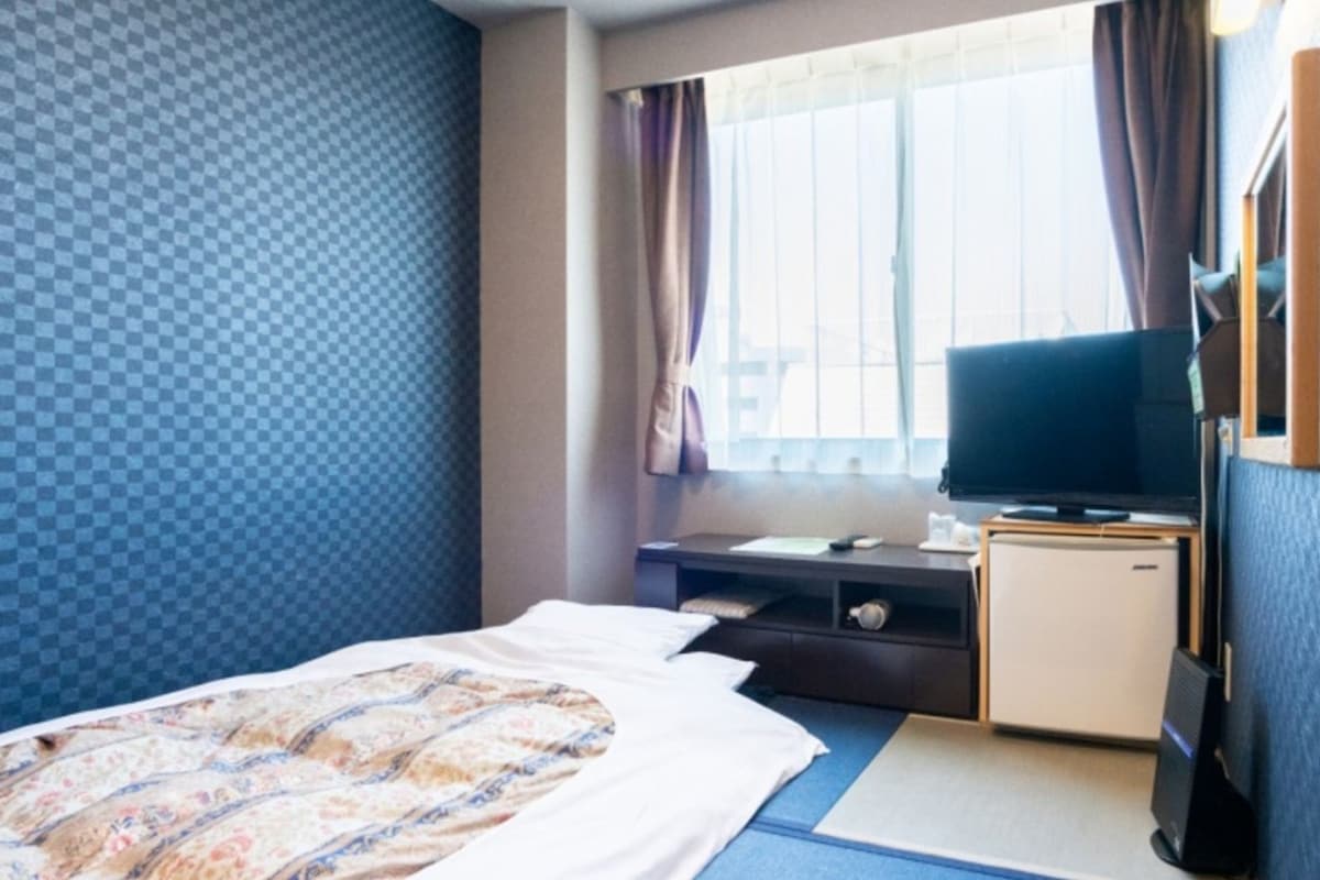 A hotel room with a bed, a desk with a television, a small refrigerator, and a window with curtains.