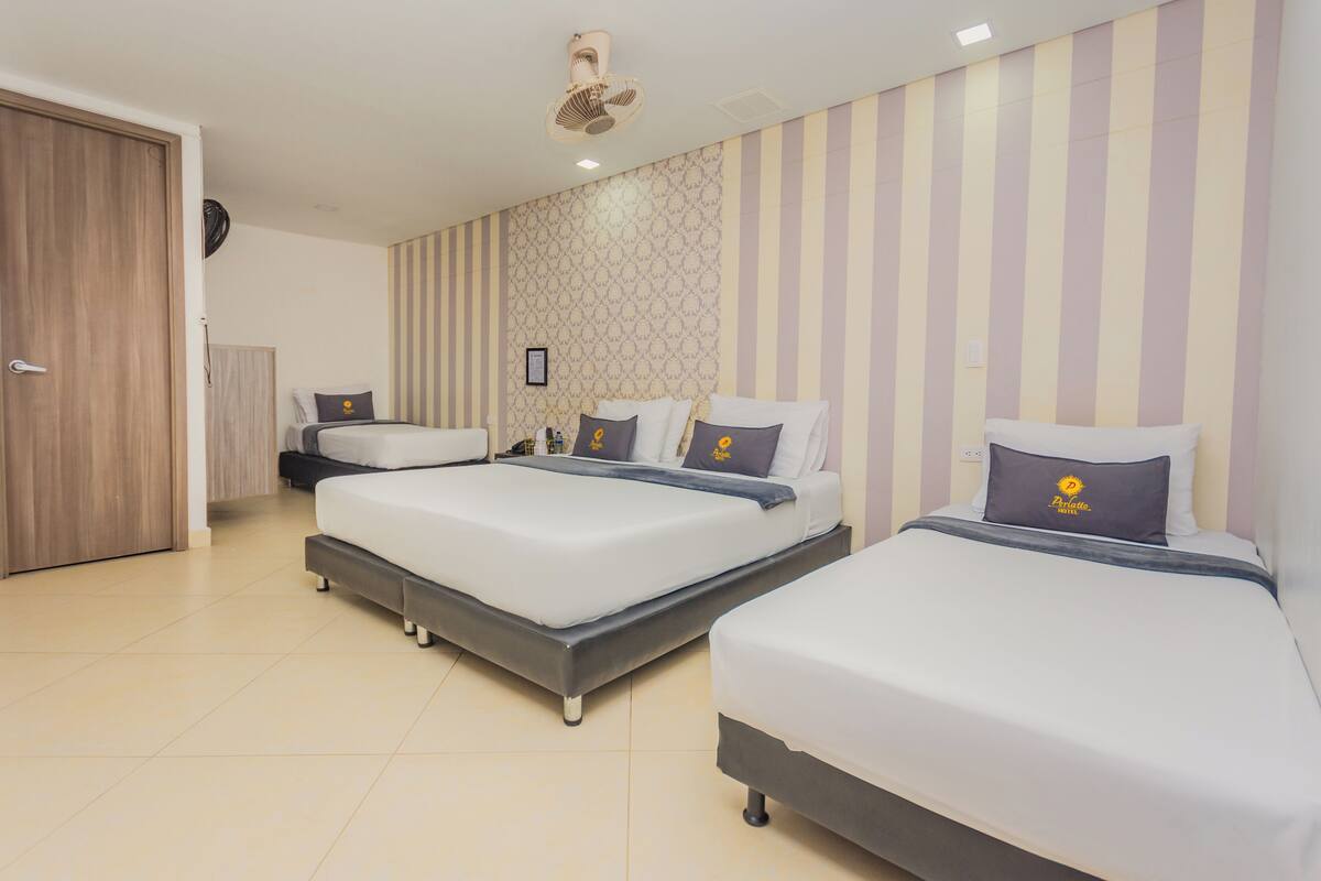Quarto de hotel com três camas, porta de madeira, ventilador de teto e papel de parede listrado.