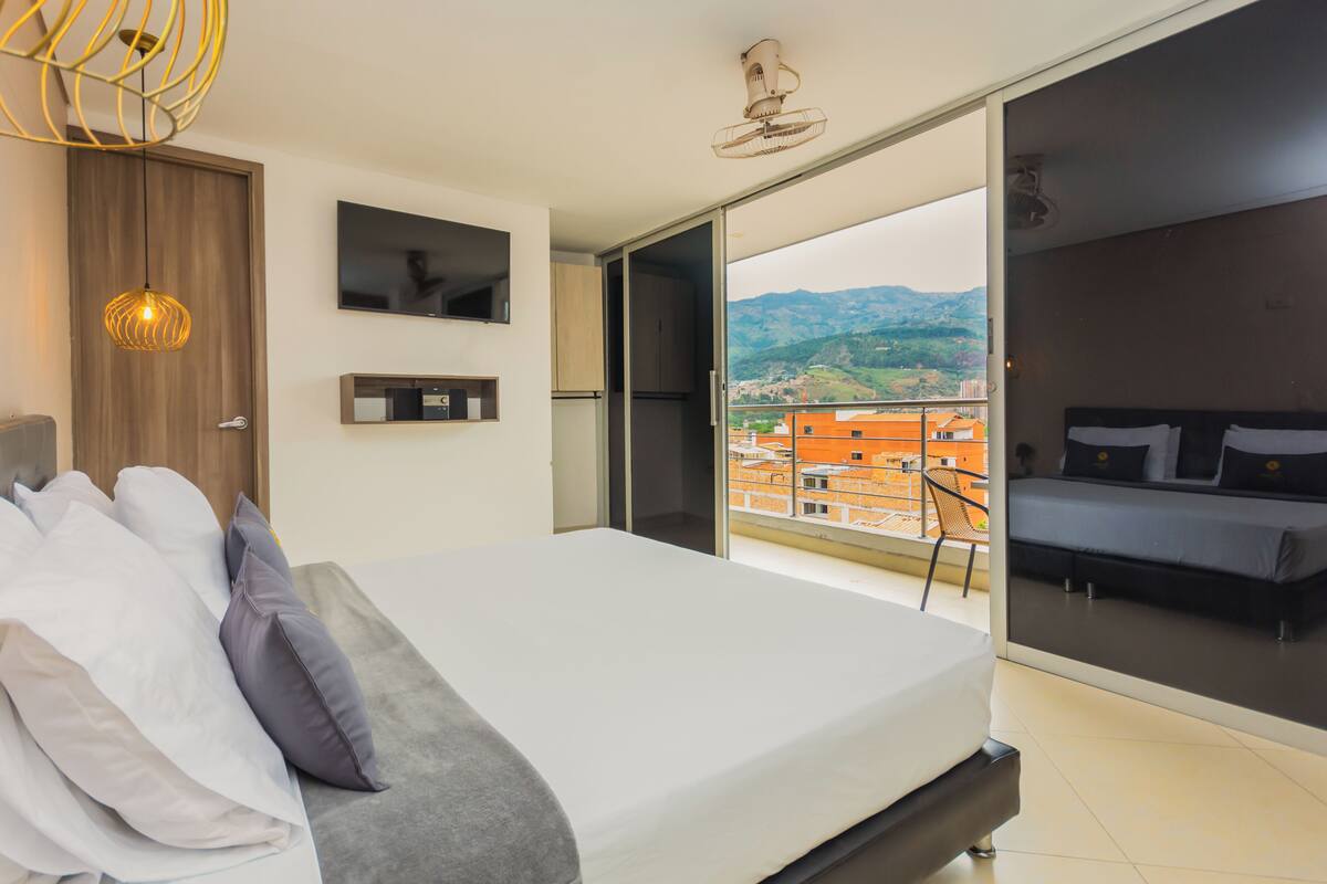 Quarto moderno com cama grande, TV e varanda com vista para a cidade.
