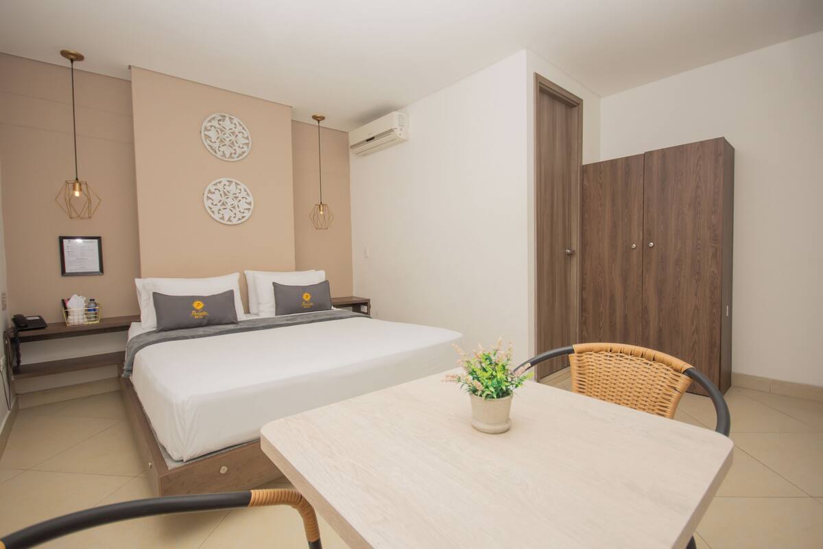 Quarto de hotel com cama, mesa de cabeceira, decoração na parede e uma mesa de jantar com uma cadeira.