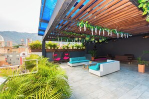 An outdoor seating area with white sofas, a wooden pergola, hanging plants, and a view of the cityscape. - Hotel Perlatto (Bello)