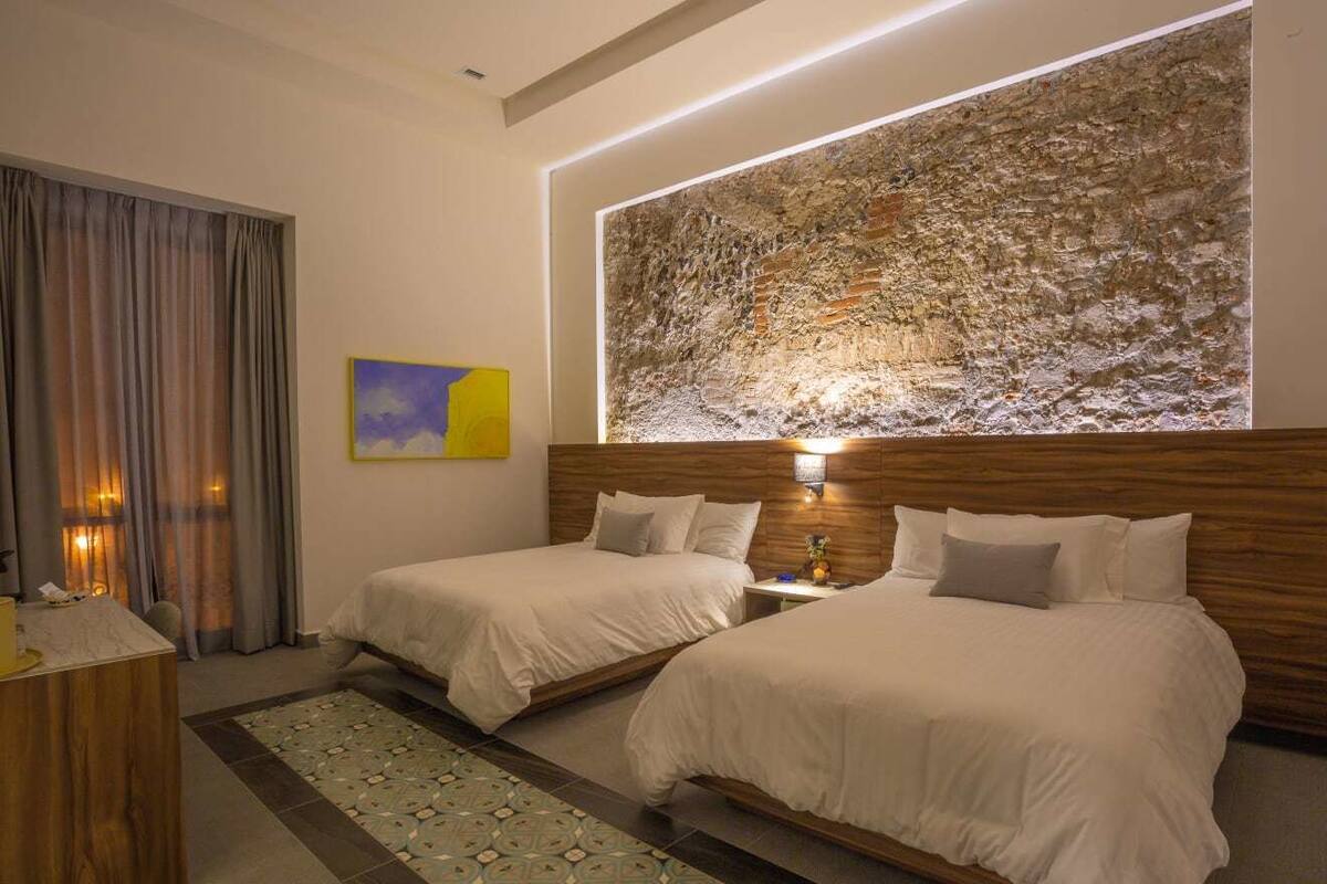 Habitación de hotel con dos camas, un cuadro de pared con textura grande, una mesita de noche con lámpara y una alfombra estampada.
