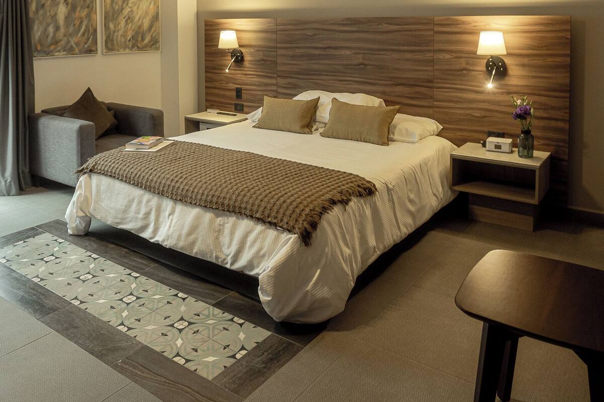 Una habitación de hotel moderna con una cama grande, dos mesitas de noche, un revistero, una silla y una alfombra decorativa.